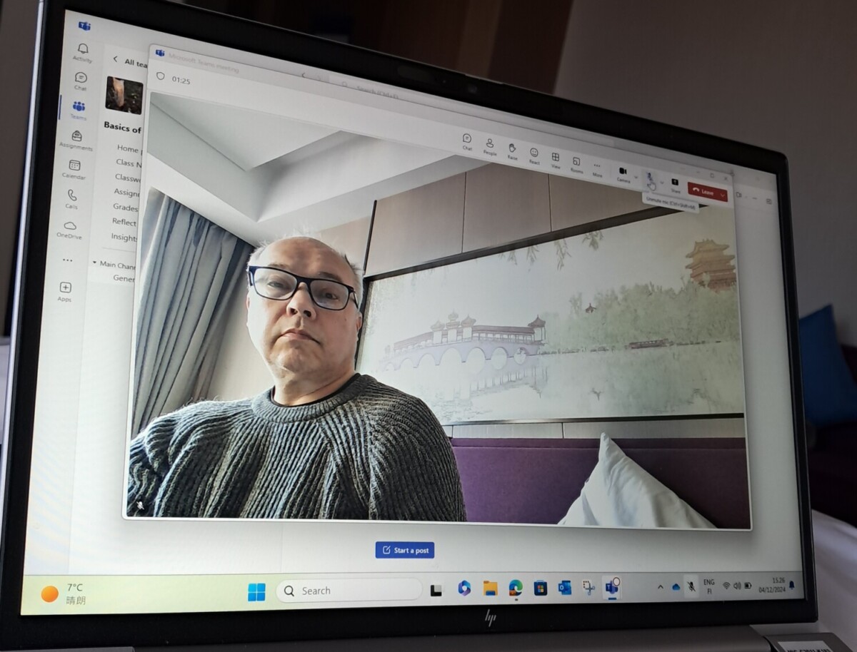 Kannettavalla tietokoneella näkyy videopuhelun näyttö, jossa on silmälaseja ja harmaata villapaitaa käyttävä henkilö, joka istuu sisätiloissa maisemamaalauksen edessä. Näkyvissä ovat myös näppäimistö ja osa huoneesta.