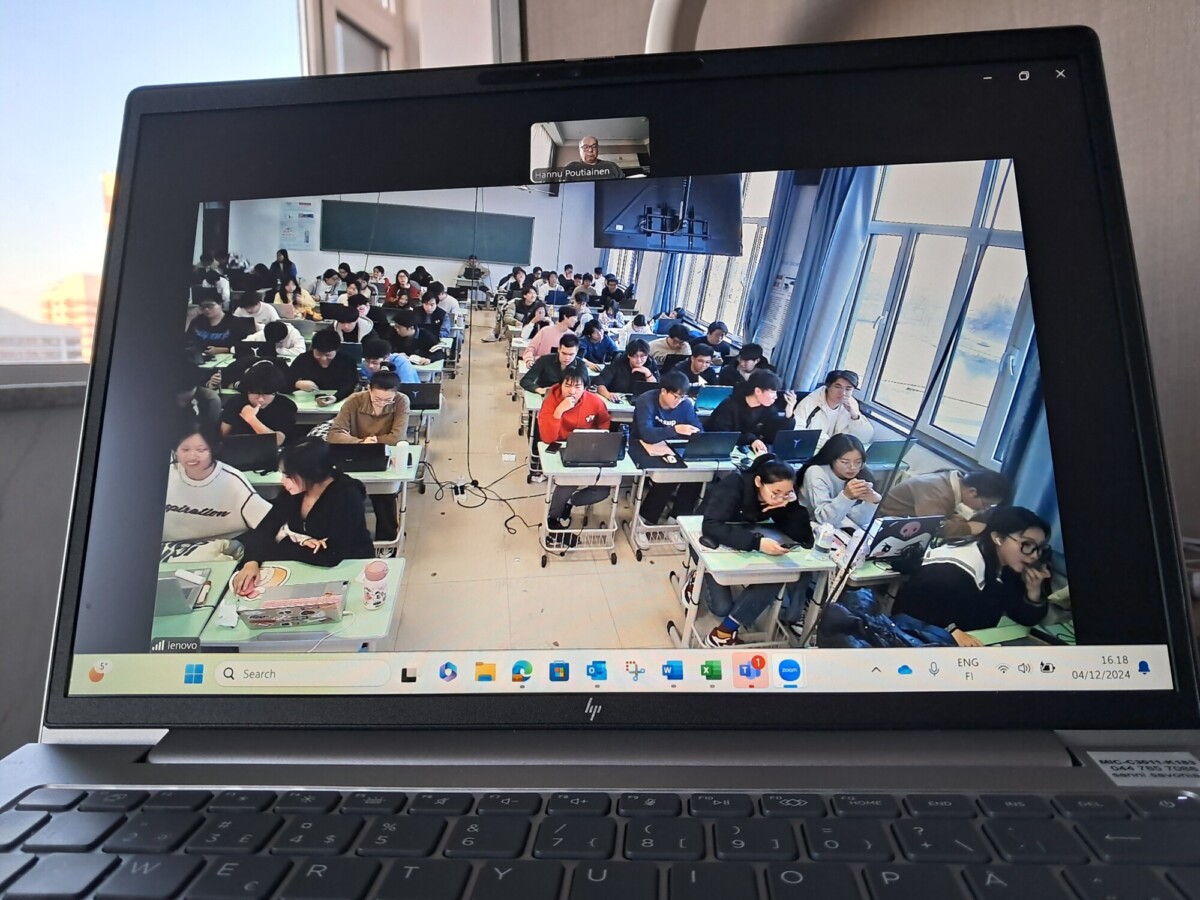 Kannettavan tietokoneen näytöllä näkyy videopuhelu, jossa luokkahuone on täynnä oppilaita, jotka istuvat pulpeteillaan, ja näytön yläosassa keskellä näkyy pieni ikkuna, jossa on yksi henkilö.