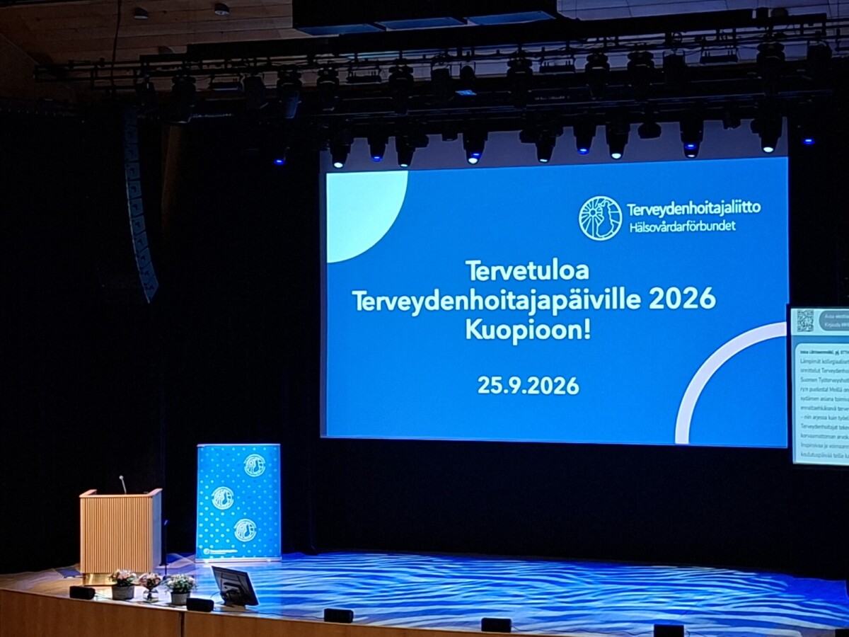 Suuri näyttämö, jonka näytöllä näkyy suomenkielinen teksti, joka toivottaa osallistujat tervetulleiksi vuonna 2026 Kuopiossa järjestettävään terveyskonferenssiin. Vasemmalla puolella on puhujalava, jossa on sininen kyltti, ja näyttämöä valaisevat ylävalot.