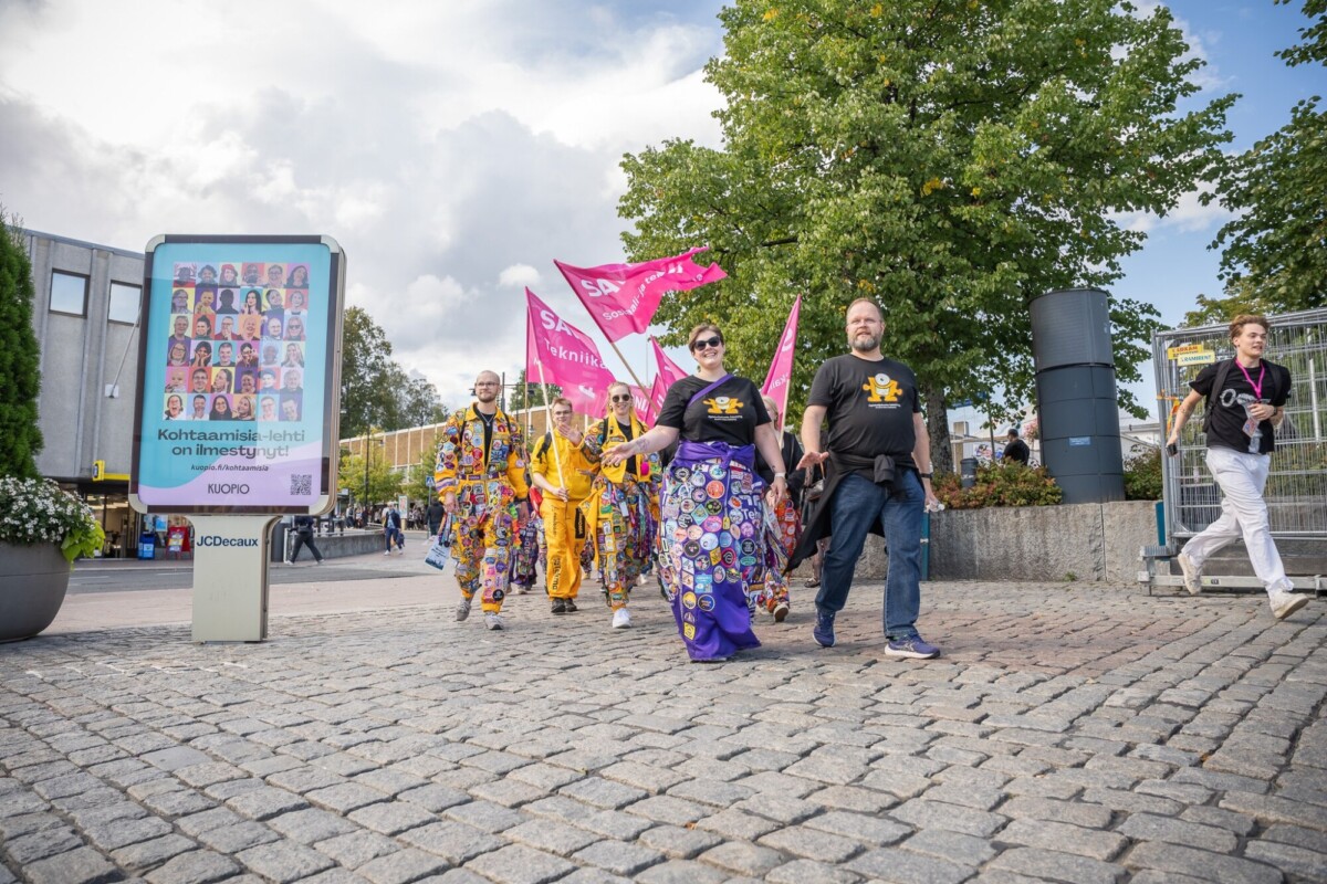 Ihmiset kävelevät ulkona mukulakivipolulla; osa heistä on pukeutunut värikkäisiin, kuviollisiin vaatteisiin ja kantaa vaaleanpunaisia lippuja. Toiset ovat pukeutuneet mustiin paitoihin, joissa on keltainen symboli. Taustalla näkyy puita ja rakennuksia.