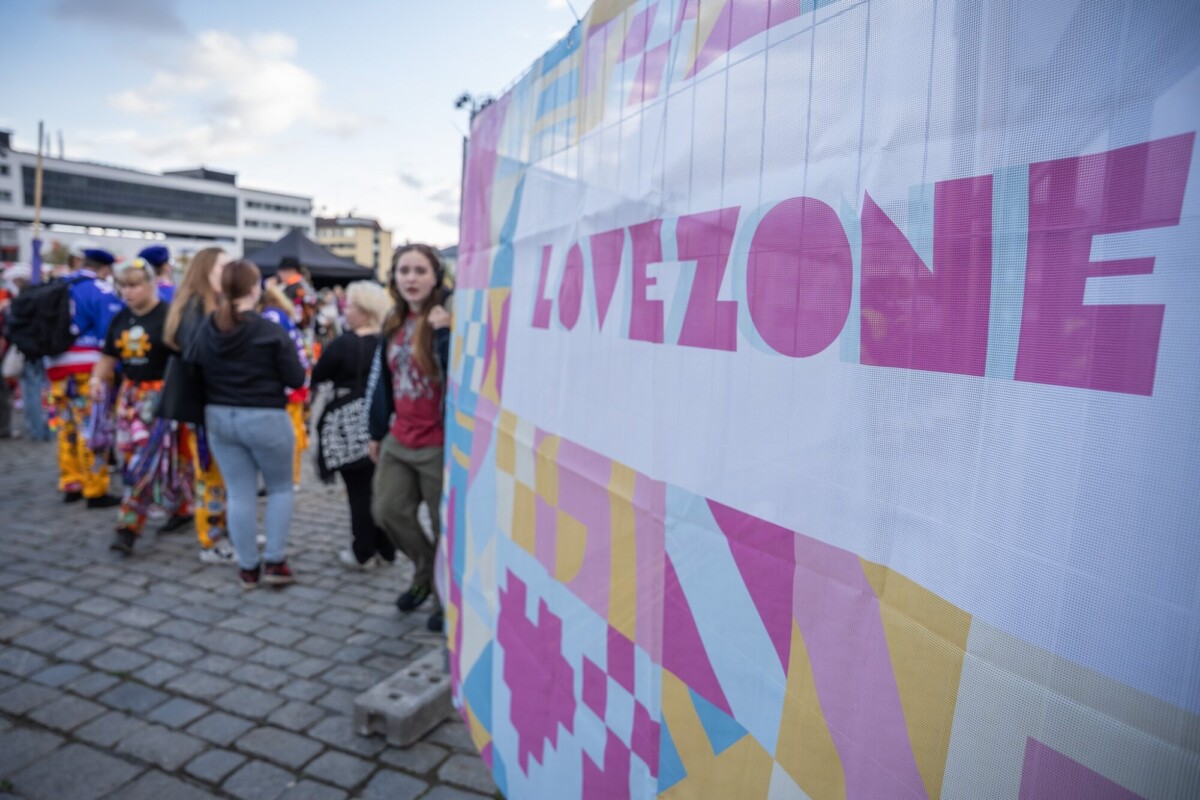 Ryhmä ihmisiä kävelee ohi värikkään banderollin, johon on painettu sana LOVEZONE, ulkoilmatapahtumassa mukulakivikadulla.