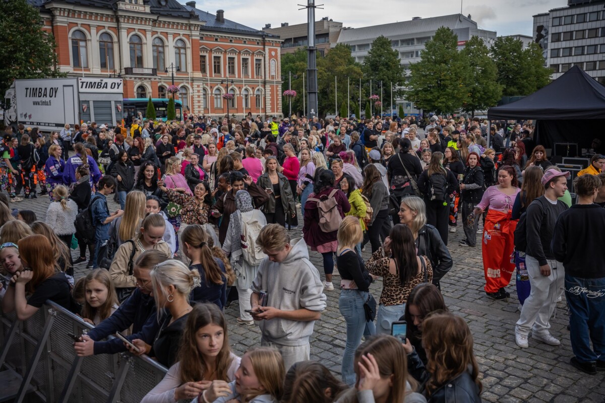 Suuri joukko ihmisiä, joukossa monia teini-ikäisiä, kokoontuu ulkona puiden ja rakennusten ympäröimälle kaupungin aukiolle julkisen tapahtuman aikana. Osa ihmisistä katselee puhelimiaan, kun taas toiset keskustelevat tai seuraavat näyttämöä.