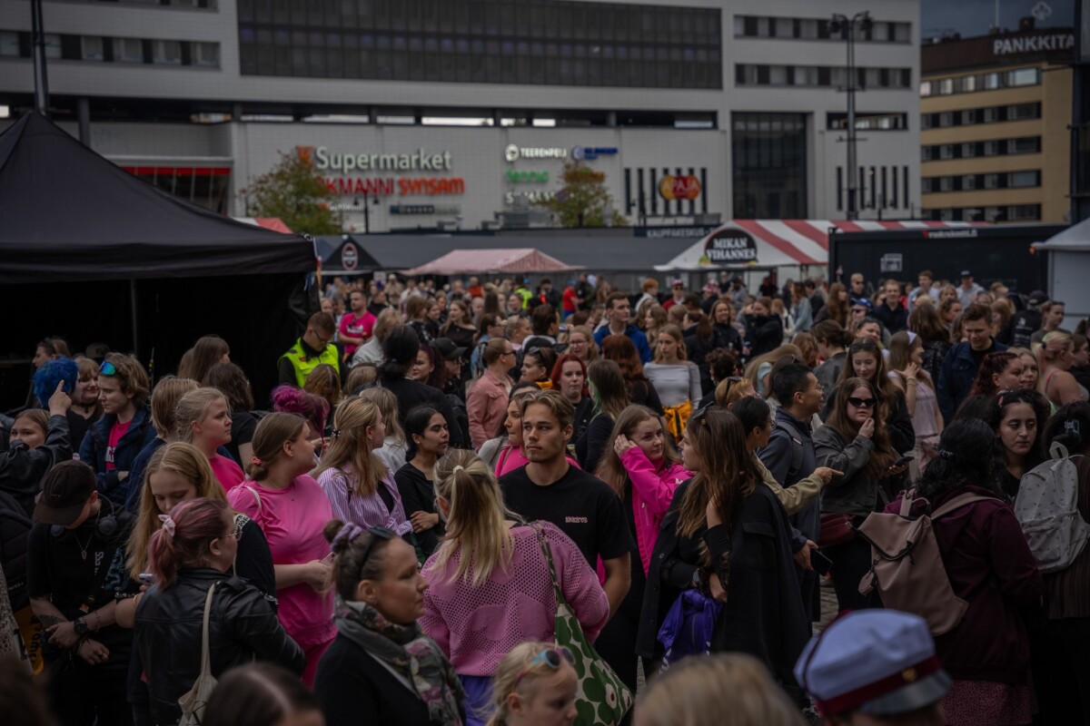 Suuri ihmisjoukko on kokoontunut ulkona kaupunkialueella supermarketin läheisyydessä. Suurin osa ihmisistä on pukeutunut rennosti, ja joillakin on yllään vaaleanpunaiset paidat. Taustalla näkyy rakennuksia ja telttoja.