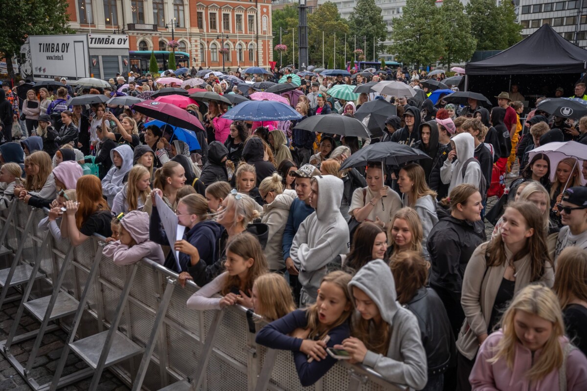 Suuri väkijoukko, josta monet pitävät sateenvarjoja kädessään, on kokoontunut ulkoilmalavan eteen sateisena päivänä. Ihmiset ovat pukeutuneet vapaa-ajan vaatteisiin ja takkeihin, ja he seisovat metalliaitojen takana; taustalla näkyy rakennuksia ja puita.
