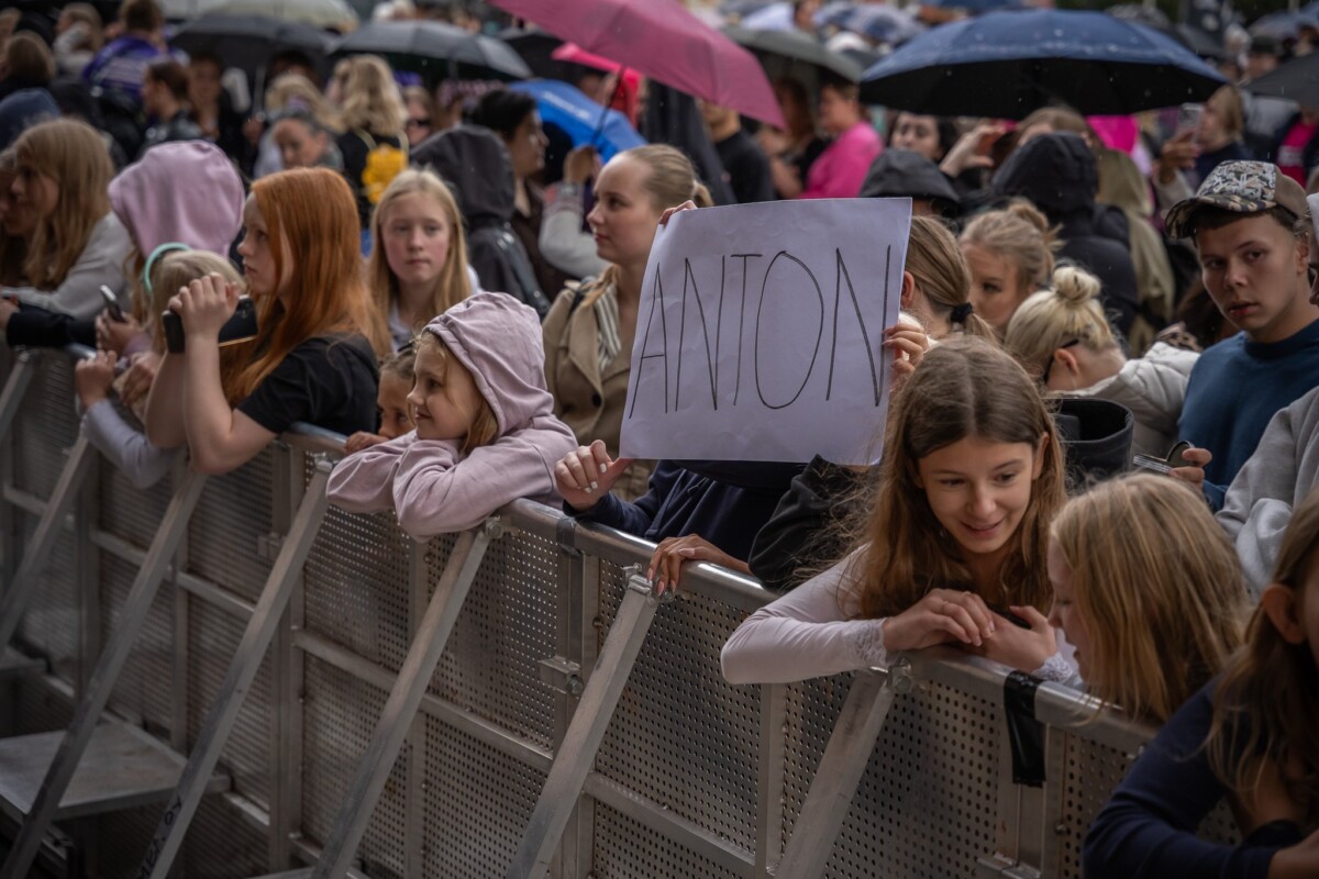 Ihmisjoukko seisoo metalliaidan takana sateessa järjestettävässä ulkoilmatapahtumassa. Osalla on sateenvarjoja, ja yksi henkilö pitää kädessään suurta kylttiä, jossa lukee ANTON. Ihmiset näyttävät innostuneilta ja keskustelevat keskenään.