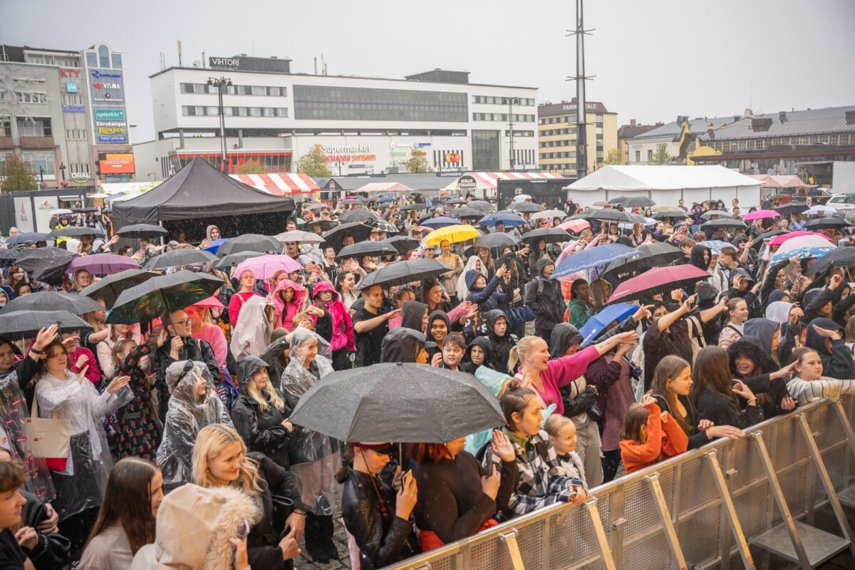 Ulkotapahtumassa suuri väkijoukko seisoo sateenvarjojen ja sadetakkien suojassa sateessa, kasvot kohti näyttämöä, ja taustalla näkyy rakennuksia ja telttoja.