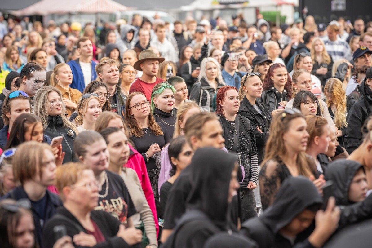 Suuri joukko ihmisiä, joista monet ovat pukeutuneet tummiin vaatteisiin, seisoo ulkona tiiviisti yhdessä ja seuraa tarkkaavaisesti jotain kuvan ulkopuolella, mikä viittaa konsertti- tai festivaalitilanteeseen.