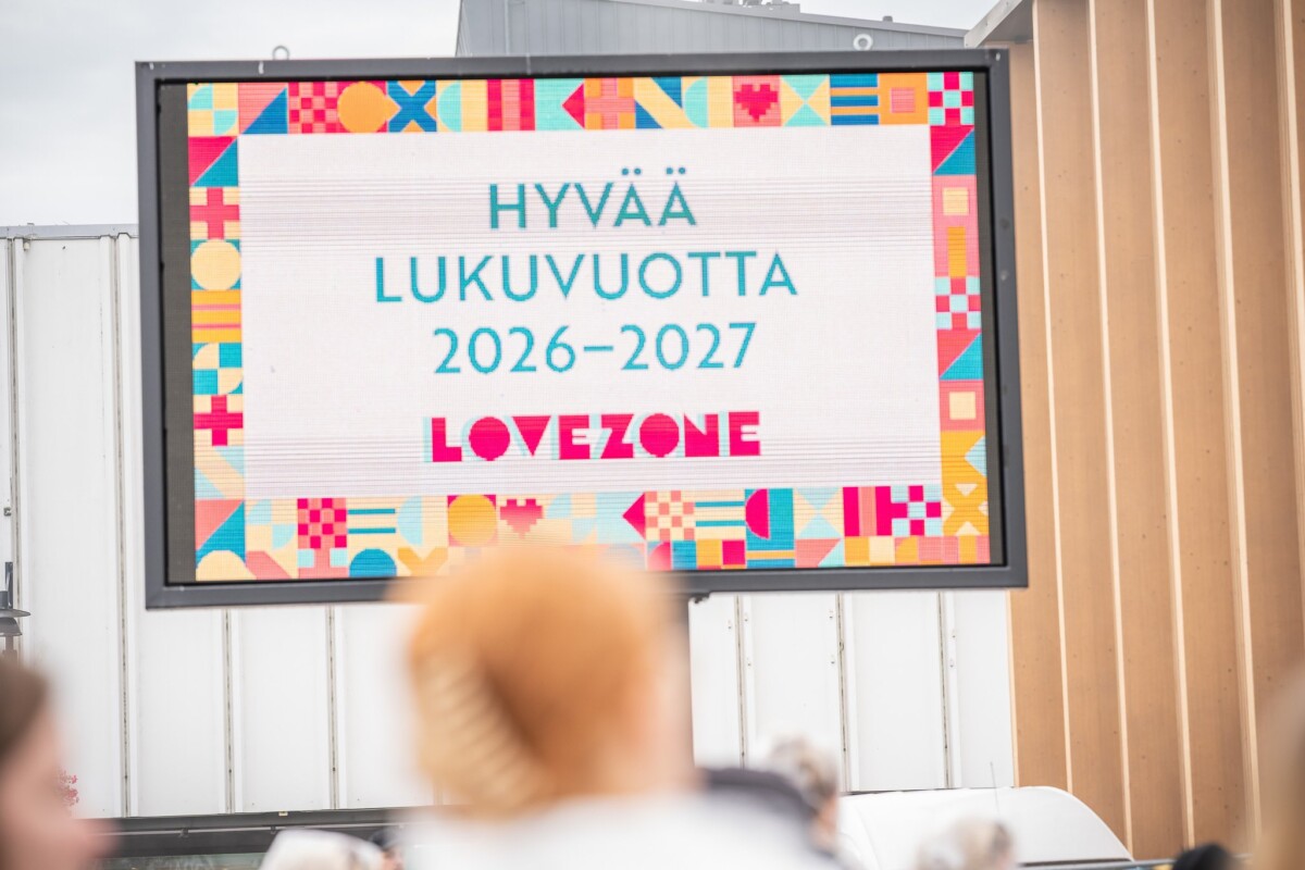 Digitaalisessa näytössä lukee ”HYVÄÄ LUKUVUOTTA 2026–2027 LOVEZONE”, ja sen ympärillä on värikkäitä geometrisia reunoja. Etualalla näkyy joitakin ihmisiä, jotka ovat hieman epäselviä.