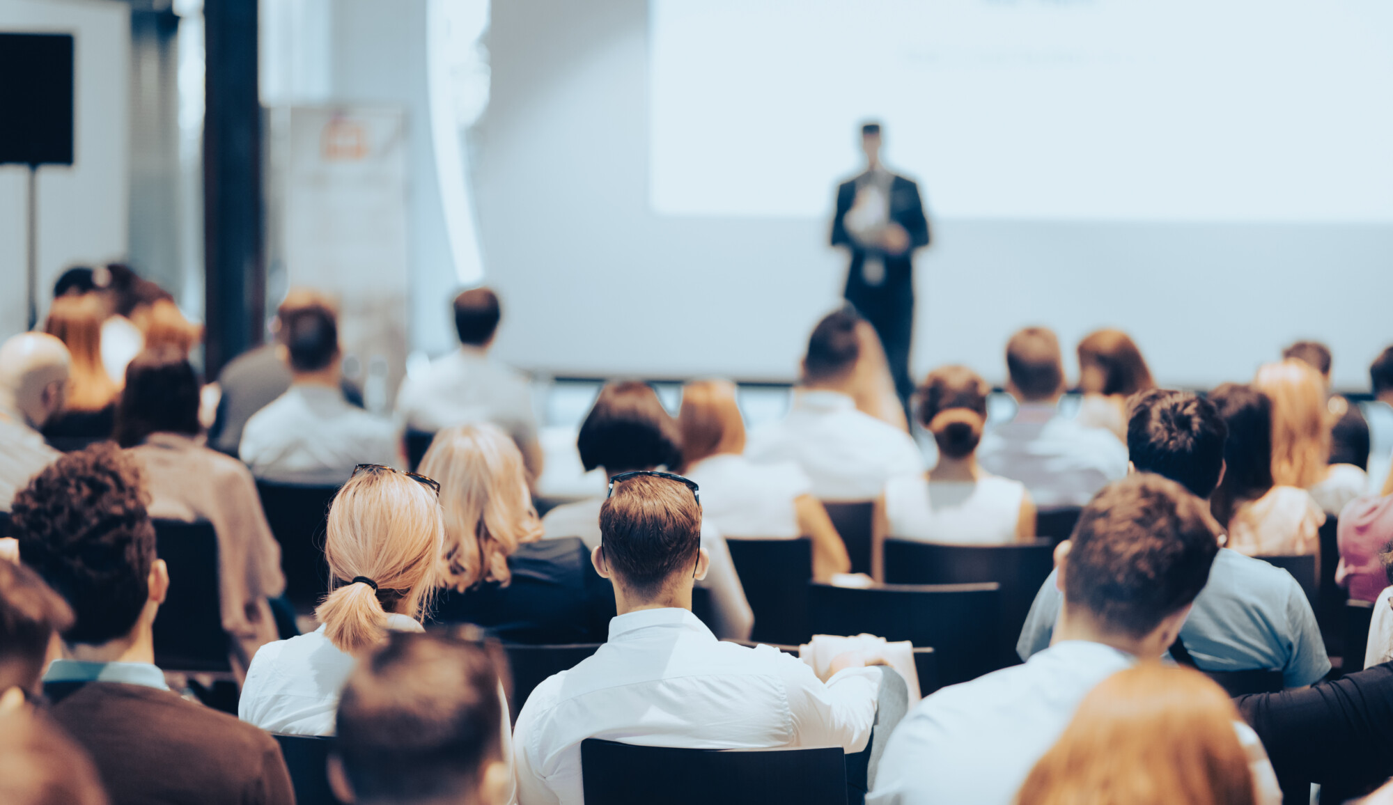 Ryhmä ihmisiä, jotka istuvat auditoriossa ja katsovat esityksen tai konferenssin aikana suurta valkokangasta vastapäätä seisovaa puhujaa.