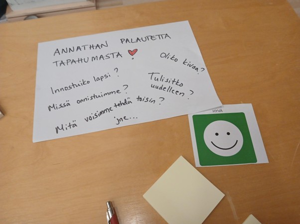 Puupöydällä on paperi, johon on kirjoitettu suomalaisia palautekysymyksiä, kynä, tyhjiä tarralappuja sekä kortti, jossa on vihreä hymyilevä kasvot ja teksti ”HYVÄ”.