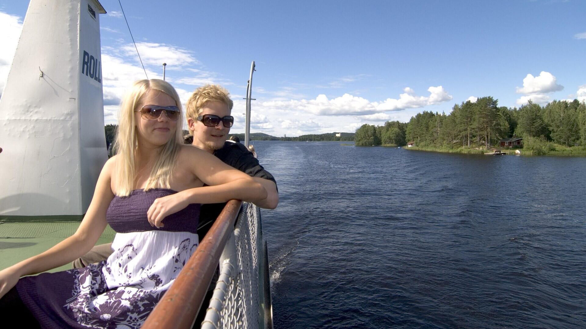 Kaksi aurinkolaseja käyttävää henkilöä seisoo lautan kannella kaiteen vieressä ja katselee rauhallista jokea, jonka rannat ovat metsäisiä, sinisen taivaan alla, jossa on hajanaisia pilviä.