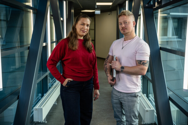 Kaksi ihmistä seisoo vierekkäin modernissa eteisessä, jossa on suuret ikkunat. Toisella on yllään punainen villapaita ja farkut, toisella vaaleanpunainen paita ja ruudulliset housut, ja hän pitää sylissään kannettavaa tietokonetta. Molemmat katsovat kameraan ja hymyilevät.