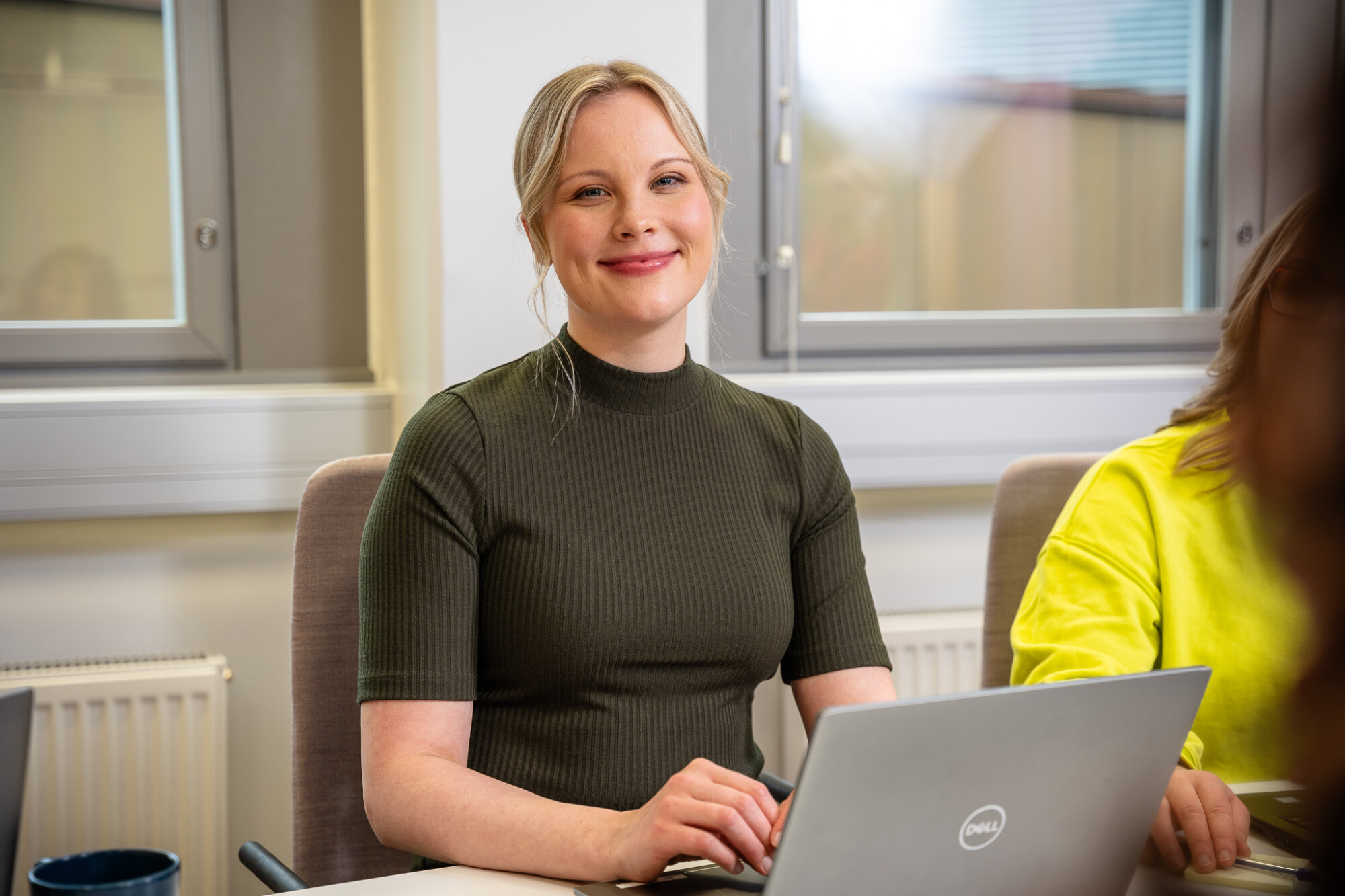Vihreää paitaa pukeutunut, hymyilevä vaaleatukkainen nainen istuu työpöydän ääressä ja käyttää Dellin kannettavaa tietokonetta. Hän on valoisassa toimistotilassa, ja lähellä on toinen henkilö, joka näkyy osittain ja jolla on yllään keltainen paita.