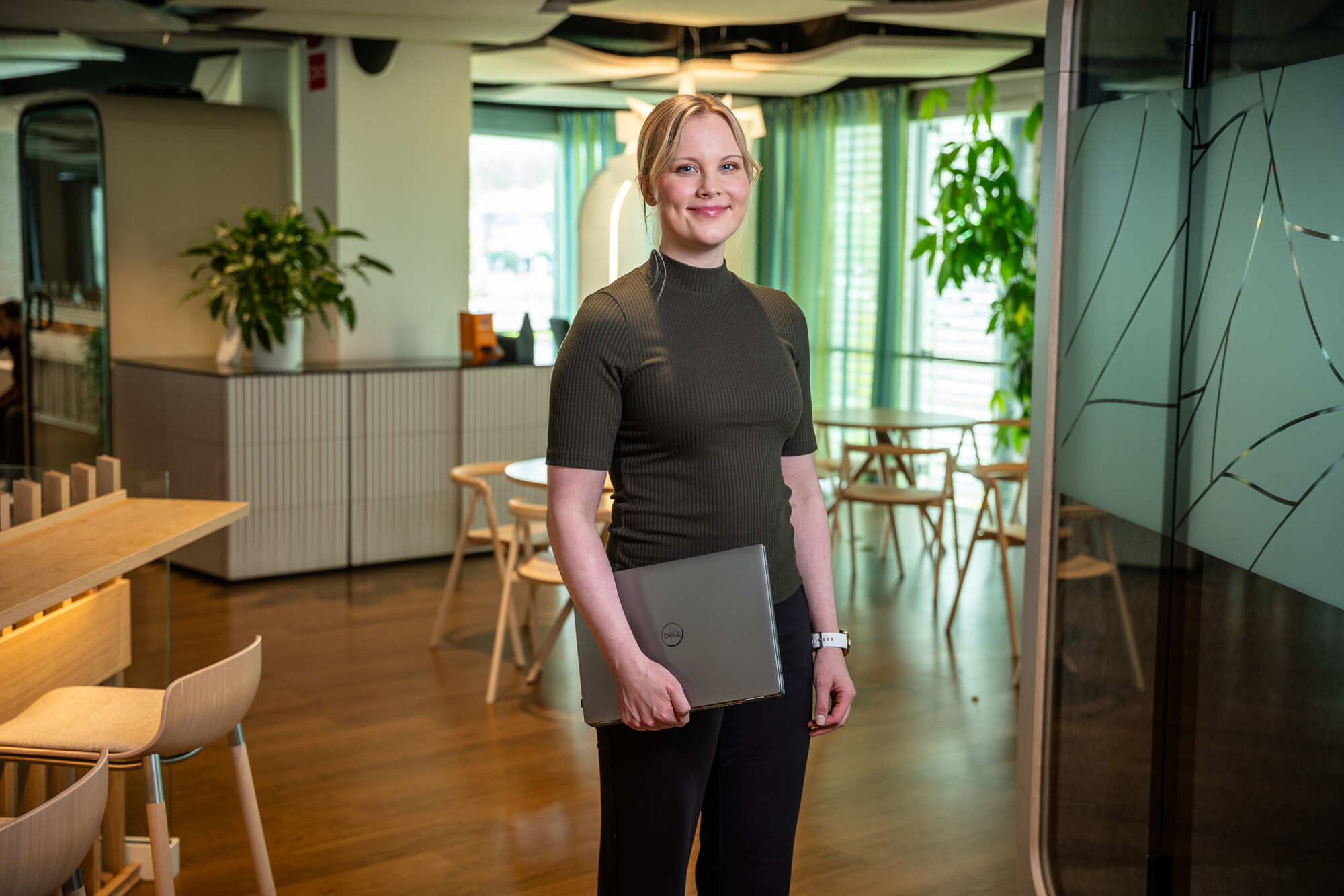 Nainen seisoo modernissa toimistossa suljetun Dell-kannettavan kanssa kädessään ja hymyilee kameraan. Toimistossa on puukalusteita, kasveja ja taustalla suuret ikkunat, joissa on vihreät verhot.