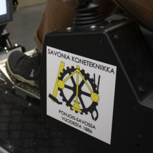 Lähikuva henkilön jalasta, joka painaa koneen poljinta. Sen vieressä on musta laatikko, jossa lukee ”Savonia Konetekniikka” sekä kelta-musta hammasrataslogo ja suomenkielistä tekstiä. Taustalla näkyy näyttö.