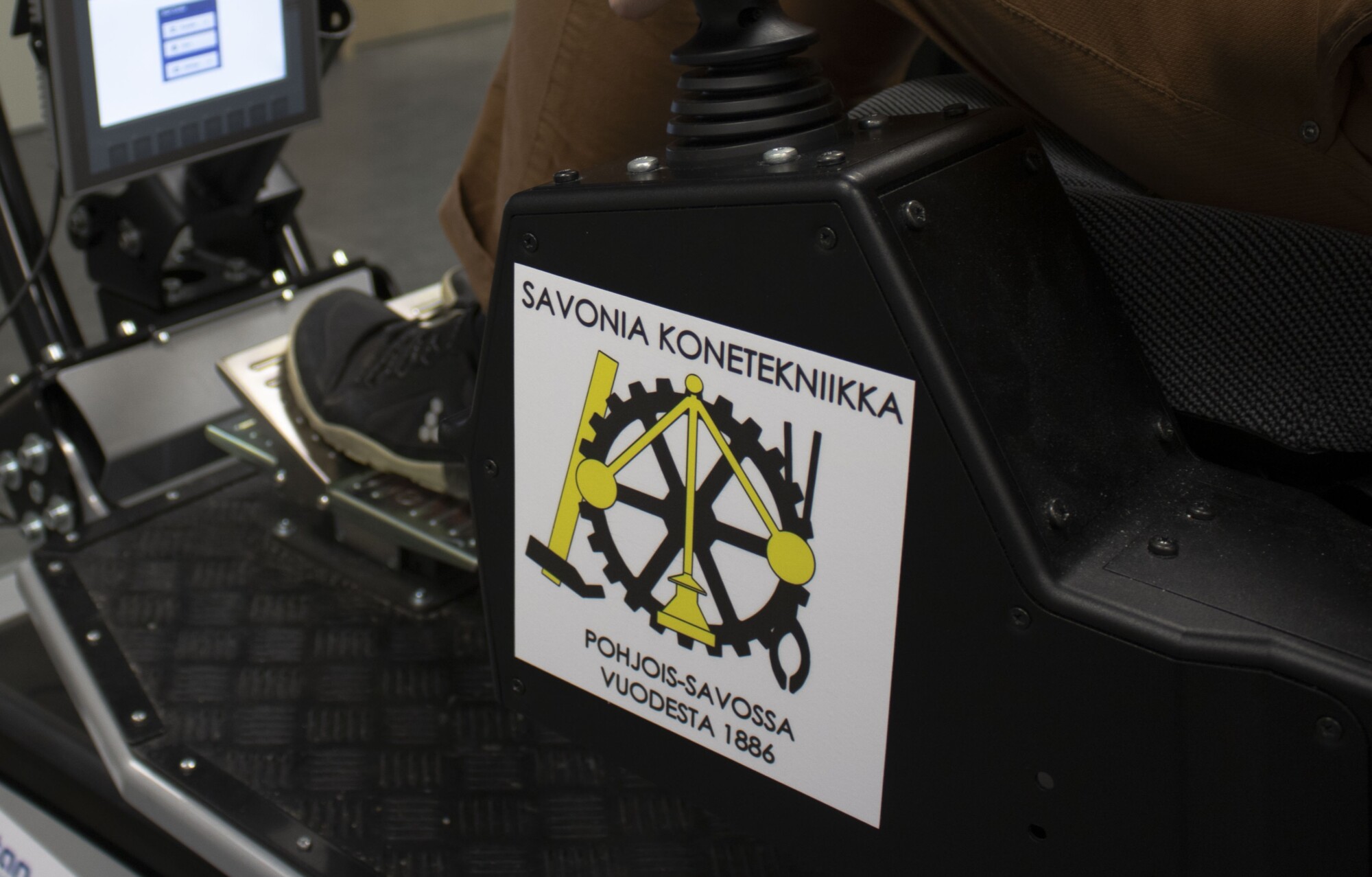 Lähikuva henkilön jalasta, joka painaa koneen poljinta. Sen vieressä on musta laatikko, jossa lukee ”Savonia Konetekniikka” sekä kelta-musta hammasrataslogo ja suomenkielistä tekstiä. Taustalla näkyy näyttö.