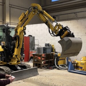 Henkilö ohjaa kaukosäätimellä keltaista Komatsu-minikaivukonetta sisätiloissa. Kaivukoneen kauha on nostettuna, ja taustalla näkyy erilaisia työkaluja ja laitteita.