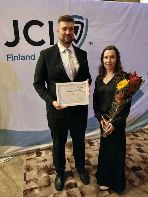 Pukumies seisoo todistuksen kanssa kädessään mustapukuista, kukkia pitelevän naisen vieressä. He poseeraavat banderollin edessä, jossa lukee ”JCI Finland”.