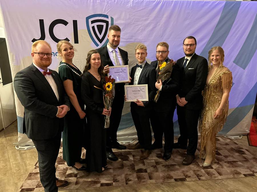 Kahdeksan juhlapukuun pukeutunutta henkilöä seisoo yhdessä tapahtumassa. Kaksi miestä keskellä pitelee todistuksia ja kukkakimppuja. Heidän takanaan on JCI-logolla varustettu banneri. Kaikki hymyilevät ja poseeraavat valokuvaa varten.