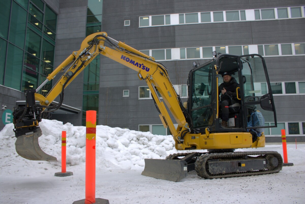 Eräs henkilö käyttää keltaista Komatsu-kaivinkonetta lumikasoiden lähellä, oranssien turvapylväiden välissä, suuren harmaan, ikkunarikkaan rakennuksen ulkopuolella.