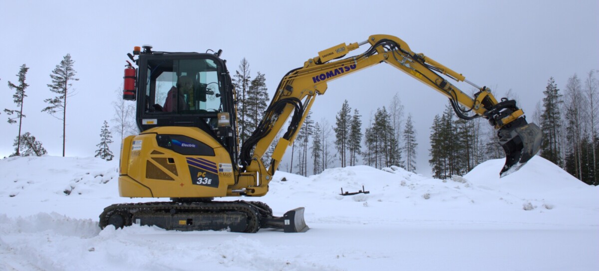 Keltainen Komatsu-minikaivinkone seisoo lumisella maalla metsän lähellä, sen puomi on ojennettuna ja kauha nostettuna. Taustalla näkyy lumipeitteisiä puita ja kukkuloita pilvisen taivaan alla.