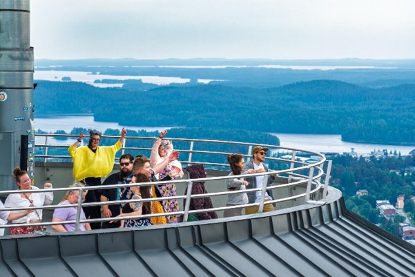 Ryhmä ihmisiä seisoo ja poseeraa tornin huipulla sijaitsevalla ulkoilmanäkötasanteella, jota ympäröivät kaukana siintävät järvet, metsät ja sumuinen taivas.