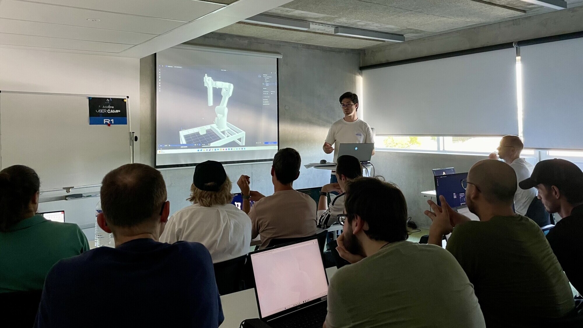 Eräs henkilö esittelee näytölle projisoitua 3D-robottikäsimallia luokkahuoneessa istuvalle, tarkkaavaiselle yleisölle, josta monet käyttävät kannettavia tietokoneita.