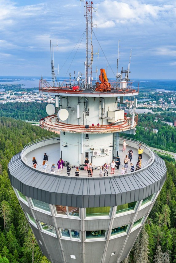 Metsän yläpuolella kohoaa korkea näkötorni, jonka huipulla on antenneja; pyöreällä näköalatasanteella kävelee ihmisiä, ja taustalla näkyy kaupunki osittain pilvisen taivaan alla.
