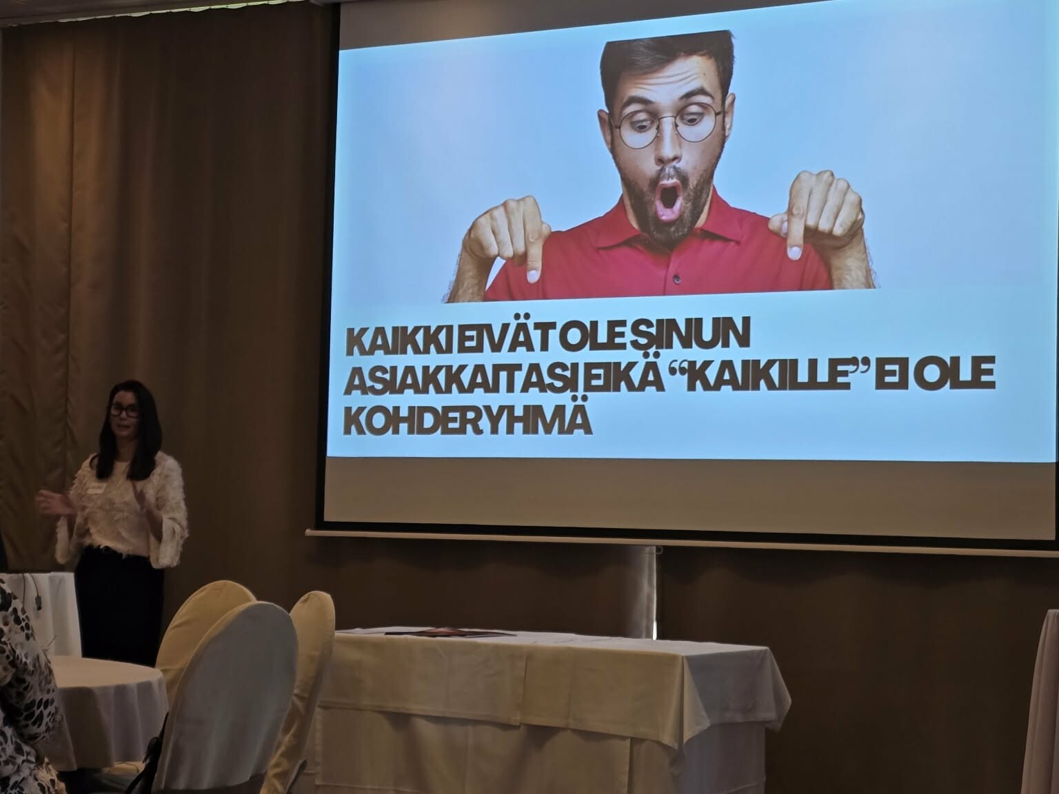 Nainen pitää esitelmää kokoushuoneessa. Näytöllä yllättynyt mies osoittaa suomenkielistä tekstiä: ”Kaikki eivät ole sinun asiakkaitasi eikä kaikki kuulu kohderyhmääsi.”.