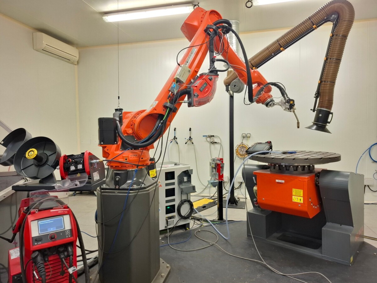 Teollinen robottivarsi on sijoitettu laboratorioon, jossa sitä ympäröivät koneet, kaapelit ja ohjausyksiköt, ja se on valmis suorittamaan automatisoituja tehtäviä, todennäköisesti valmistus- tai hitsausympäristössä.