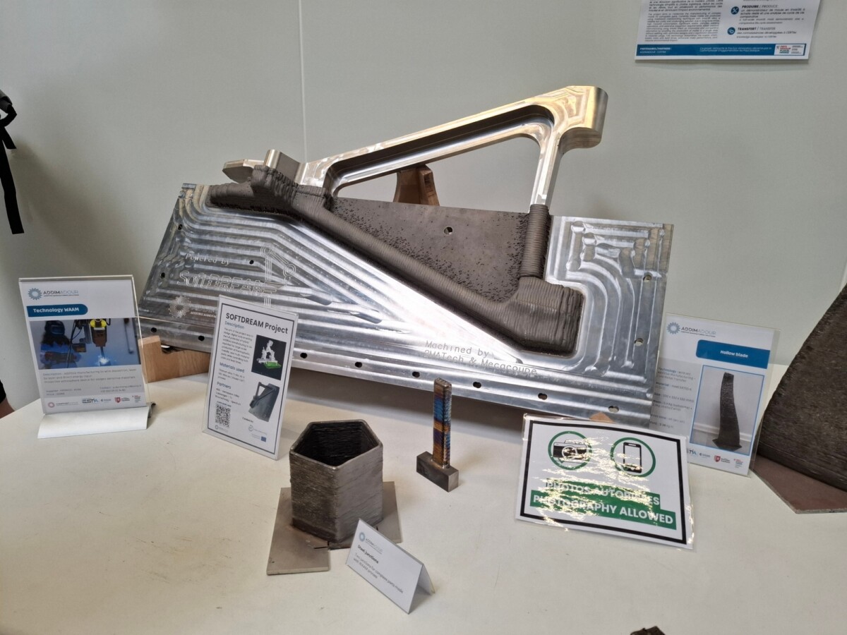 Metallinen 3D-tulostettu osa on esillä koneistetulla alustalla, ja sen ympärillä on valkoisella pöydällä opastekylttejä sekä näytteitä tulostetuista metalliesineistä. Paikalla on myös kyltti, jossa lukee ”Valokuvaus sallittu”.