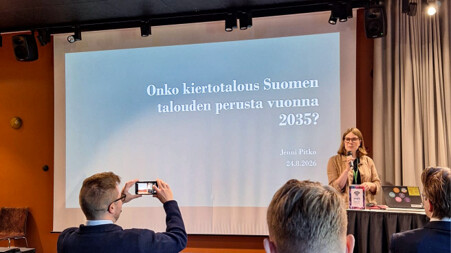 Nainen seisoo puhujakorokkeen vieressä ja pitää esitelmää suomeksi, ja yleisössä on ihmisiä. Dia-esityksessä lukee: ”Onko kiertotalous Suomen talouden perusta vuonna 2035?”.