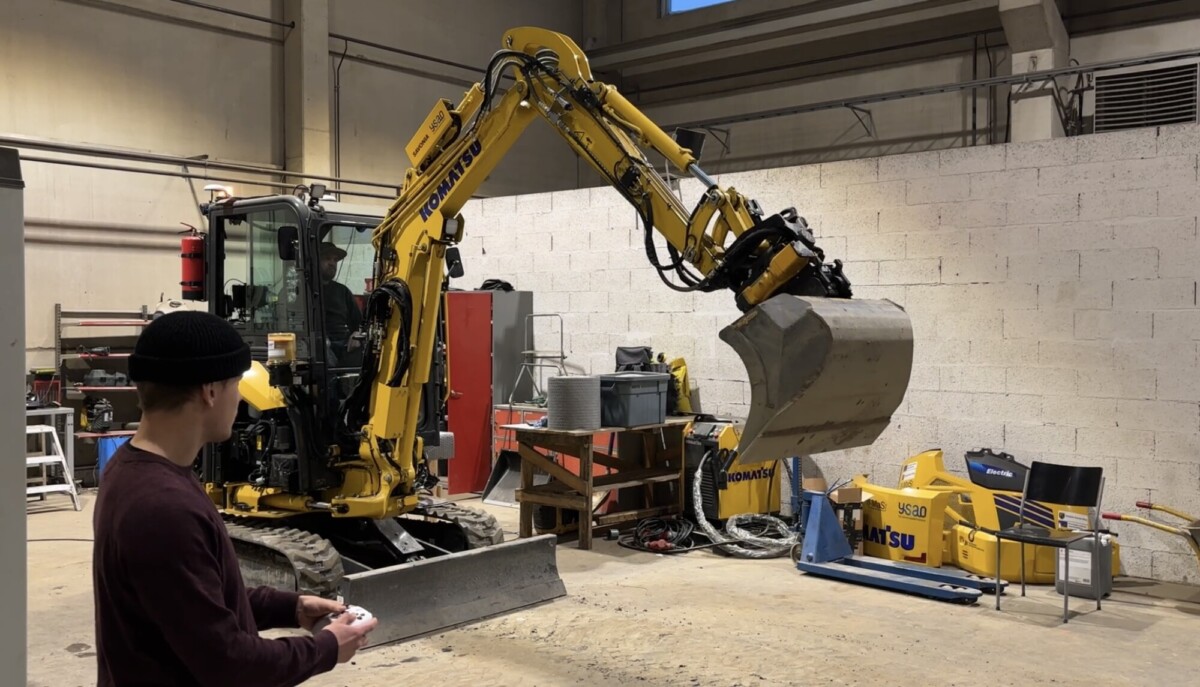 Eräs henkilö ohjaa kaukosäätimellä keltaista Komatsu-minikaivukonetta sisätiloissa. Kaivukoneen kauha on nostettu ylös työpaja-alueen läheisyydessä, jossa on työkaluja ja laitteita.
