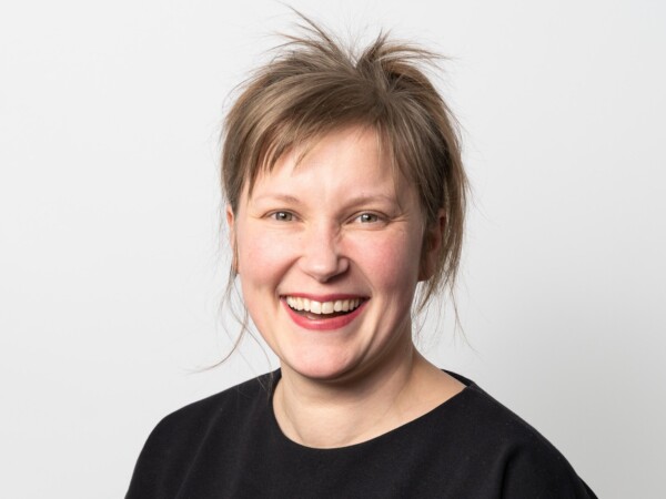 Nainen, jolla on lyhyet, pöyheät vaaleanruskeat hiukset, hymyilee leveästi kameraan. Hänellä on yllään musta paita, ja hän poseeraa yksinkertaista valkoista taustaa vasten.