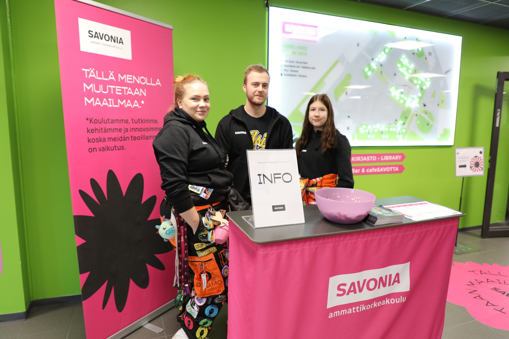 Kolme henkilöä seisoo Savonia-kyltillä varustetun infopisteen takana. Heillä on yllään värikkäät haalarit. Heidän takanaan on vaaleanpunaisia banderolleja ja karttaa näyttävä näyttö. Paikka näyttää olevan koulu tai yliopisto.