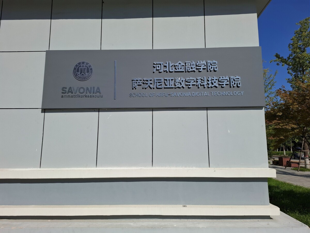 Rakennuksen kyltissä lukee SAVONIA ammattikorkeakoulu sekä SCHOOL OF HEBEI-SAVONIA DIGITAL TECHNOLOGY englanniksi, suomeksi ja kiinaksi. Taustalla näkyy puita ja polku.