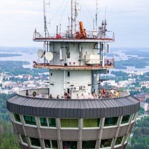 Lähikuva korkean tornin näköalatasanteelta, jossa ihmiset katselevat alla avautuvaa kaupunkia ja järviä; maisemassa näkyy rakennuksia, puita ja vettä pilvisen taivaan alla.