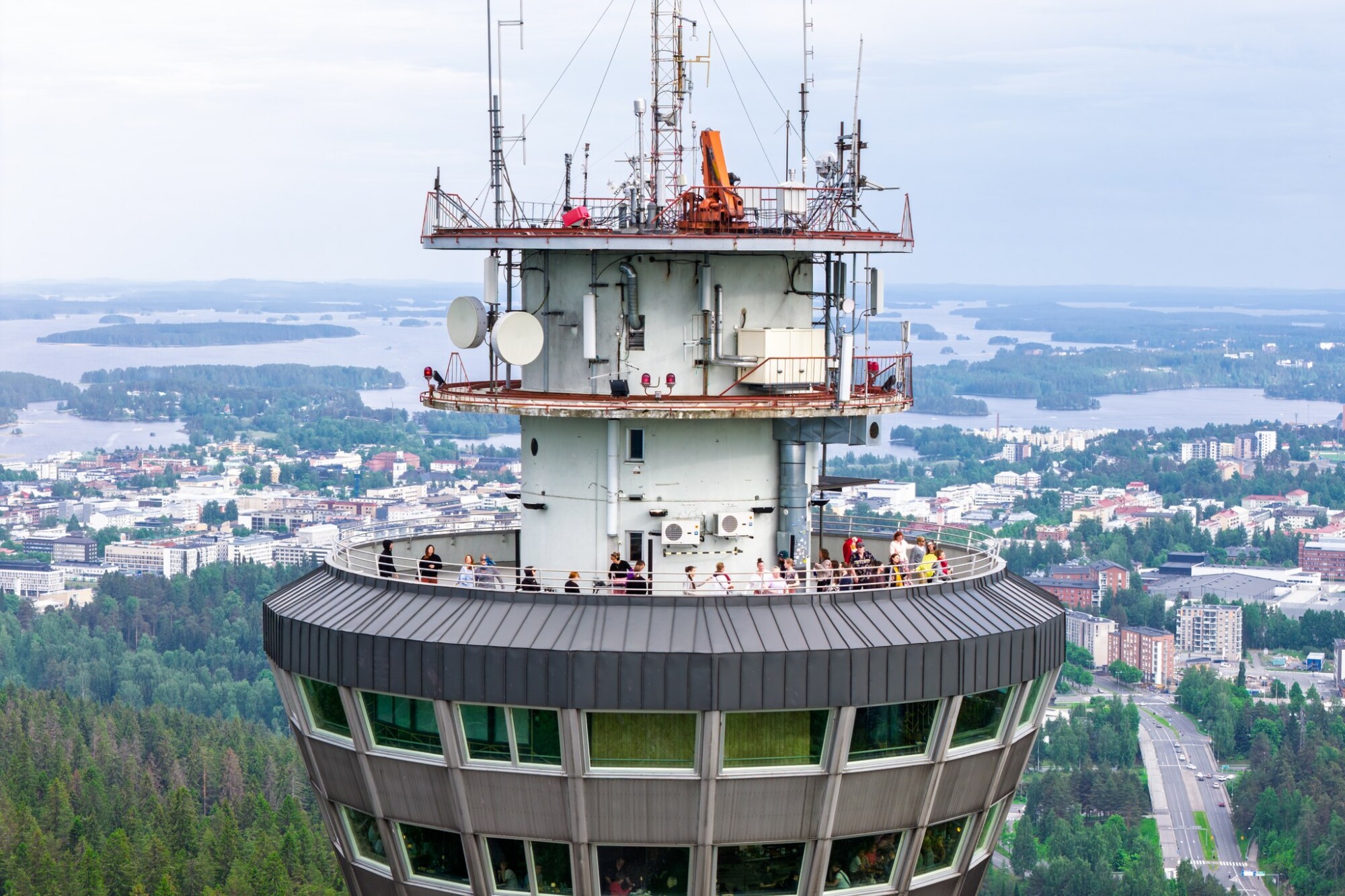 Lähikuva korkean tornin näköalatasanteelta, jossa ihmiset katselevat alla avautuvaa kaupunkia ja järviä; maisemassa näkyy rakennuksia, puita ja vettä pilvisen taivaan alla.