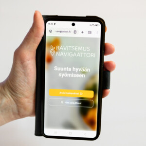 Kädessä on älypuhelin, jonka näytöllä näkyy suomalainen verkkosivusto nimeltä Ravitseus Navigaattori. Sivustolla on painike ”Arvioi ruokavaliosi” sekä toinen painike, jossa lukee ”Anna palautetta”. Tausta on sumea, ja siinä näkyy oransseja ja vihreitä värejä.