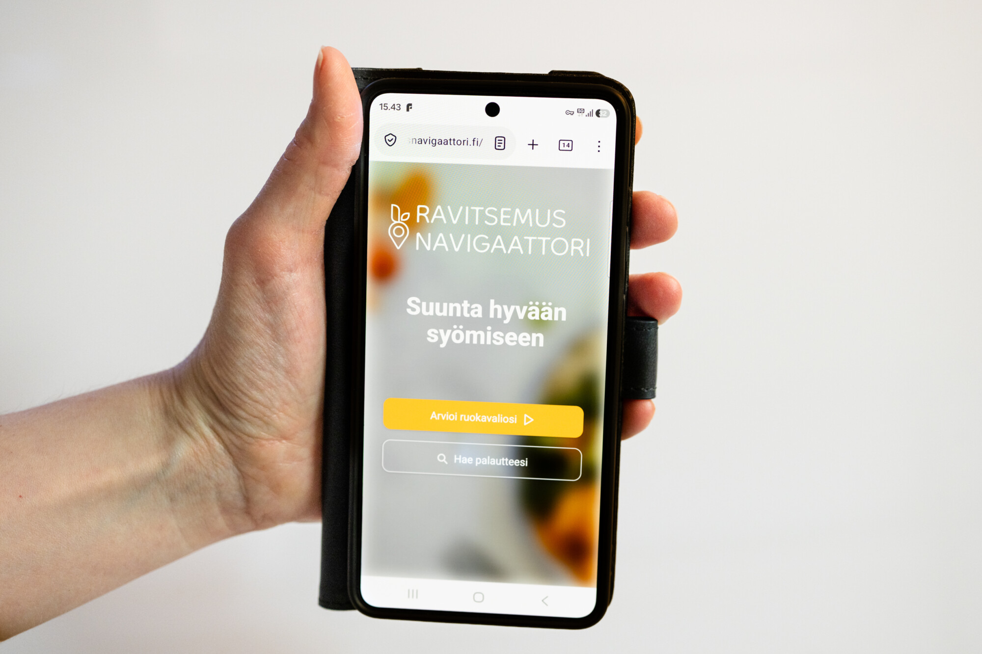 Kädessä on älypuhelin, jonka näytöllä näkyy suomalainen verkkosivusto nimeltä Ravitseus Navigaattori. Sivustolla on painike ”Arvioi ruokavaliosi” sekä toinen painike, jossa lukee ”Anna palautetta”. Tausta on sumea, ja siinä näkyy oransseja ja vihreitä värejä.