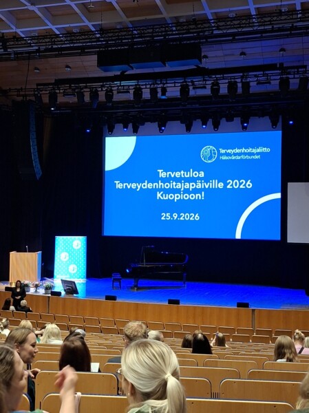 Konferenssisali, jossa ihmiset istuvat näyttämöä kohti. Suurella näytöllä lukee: ”Tervetuloa Terveydenhoitajapäiville 2026 Kuopioon! 25.9.2026.” Näyttämöllä on piano ja puhujalava.