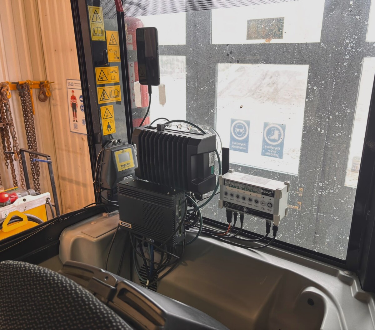 Näkymä ajoneuvon sisältä, jossa näkyvät elektroniset ohjausyksiköt, johdotus, varoitusmerkinnät sekä ikkuna, jonka ulkopuolella näkyy kylttejä ja lunta.