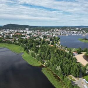 Ilmakuva järven rannalla sijaitsevasta kaupungista, jossa on reheviä vihreitä puita, hiekkarantoja ja kaukana näkyviä rakennuksia osittain pilvisen taivaan alla.