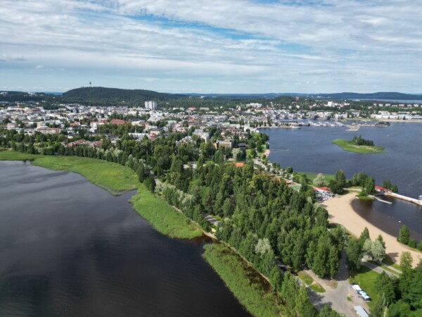 Ilmakuva järven rannalla sijaitsevasta kaupungista, jossa on reheviä vihreitä puita, hiekkarantoja ja kaukana näkyviä rakennuksia osittain pilvisen taivaan alla.