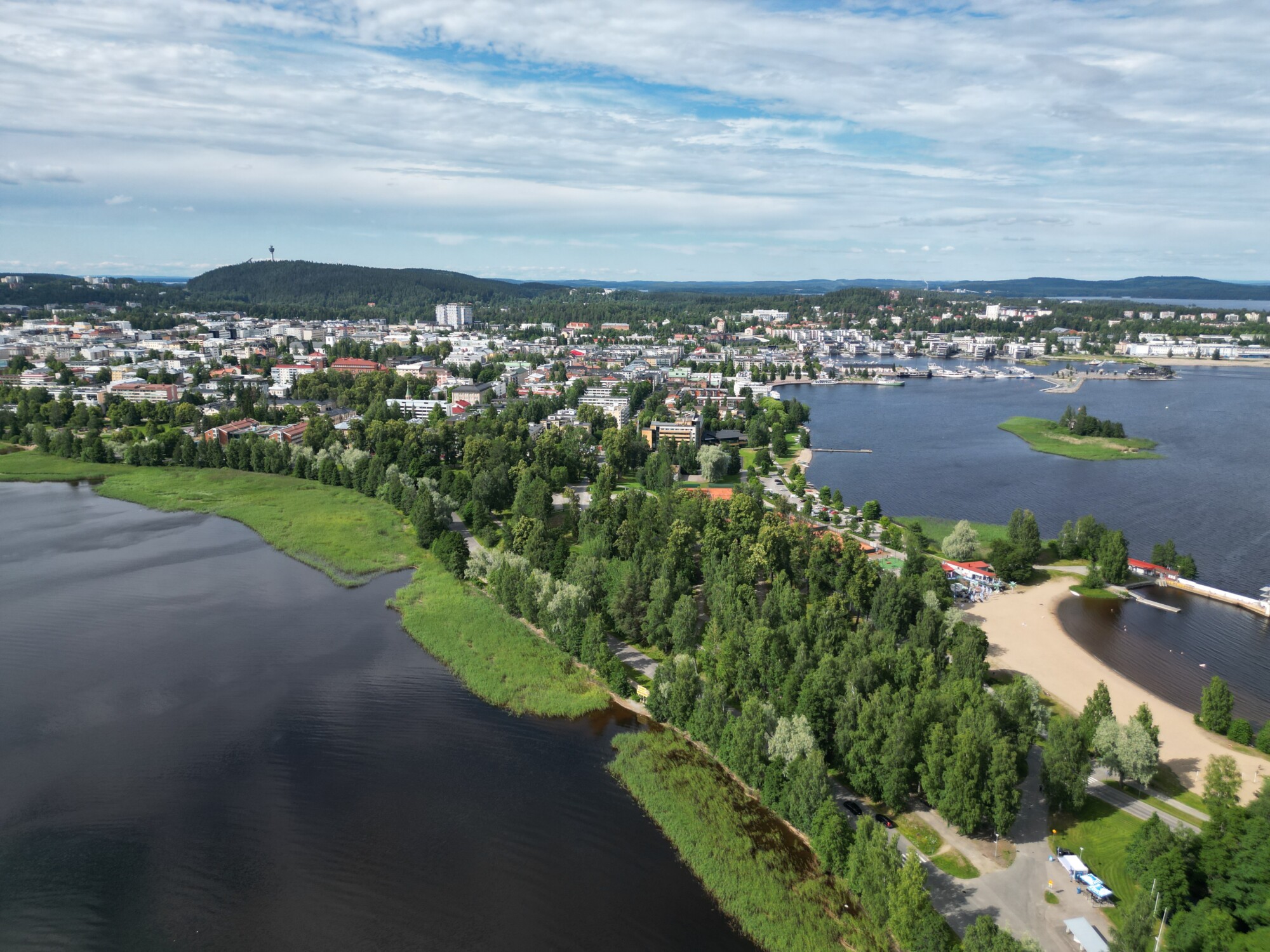 Ilmakuva järven rannalla sijaitsevasta kaupungista, jossa on reheviä vihreitä puita, hiekkarantoja ja kaukana näkyviä rakennuksia osittain pilvisen taivaan alla.