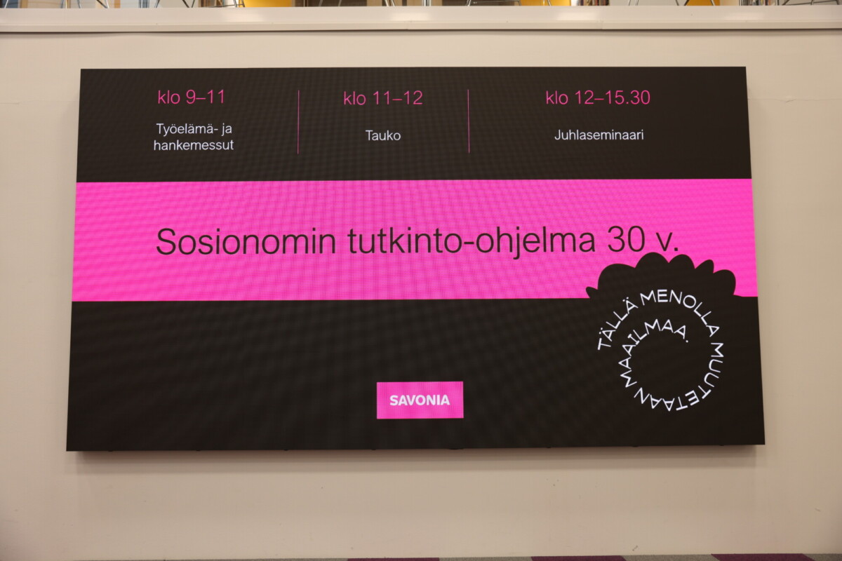 Digitaalinen näyttö, jossa näkyy Sosionomin tutkinto-ohjelman 30-vuotisjuhlatapahtuman ohjelma, jossa on tapahtumien ajat ja kuvaukset suomeksi vaaleanpunaisella ja mustalla taustalla.