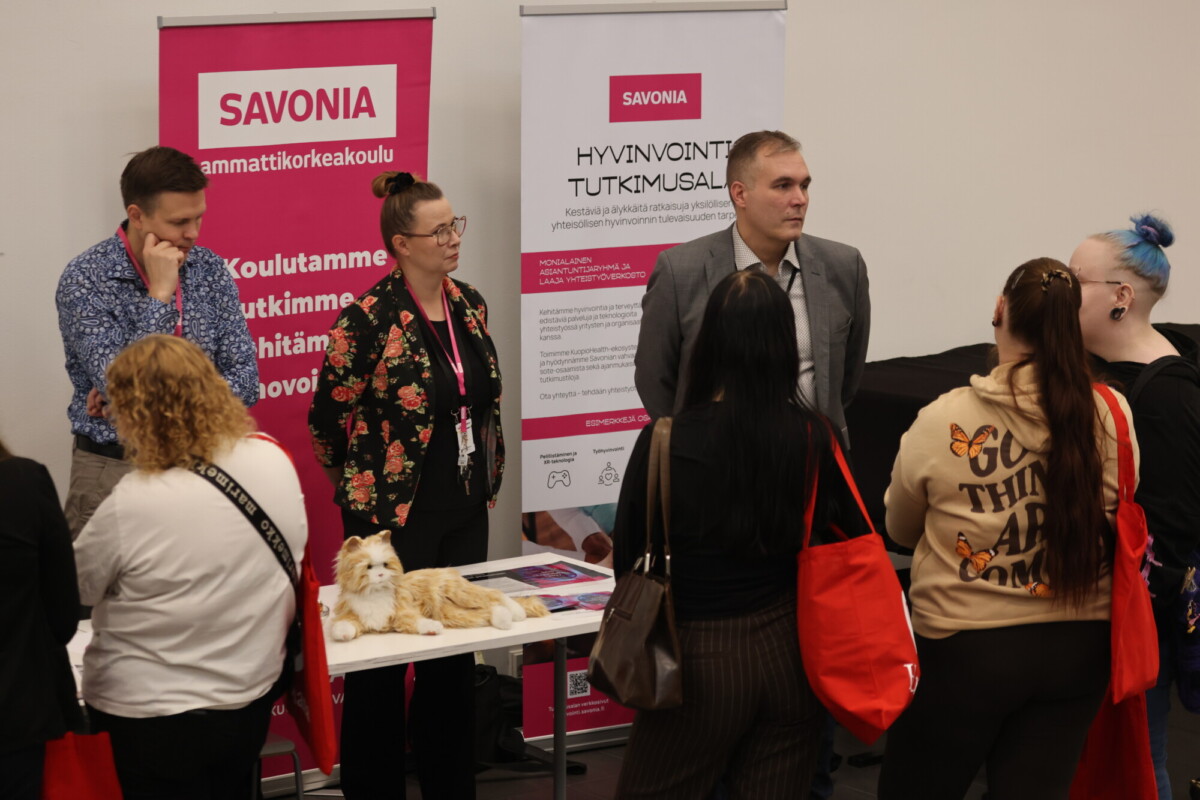 Useat ihmiset keskustelevat Savonian ammattikorkeakoulun osastolla koulutustapahtumassa. Paikalla näkyy kaksi banderollia sekä pöytä, jolla on esitteitä ja pehmolelu. Jotkut kävijät pitävät käsissään punaisia kangaskasseja ja kuuntelevat edustajia.