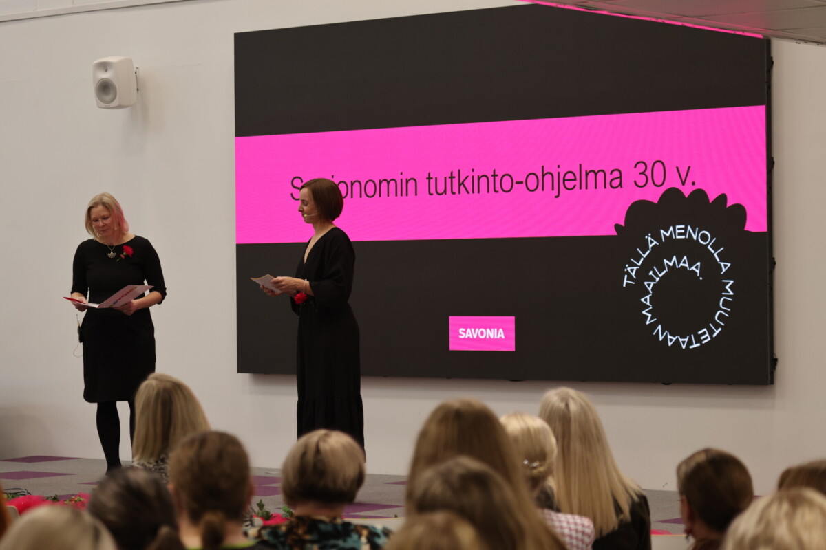 Kaksi naista seisoo lavalla suuren näytön edessä, jolla näkyy suomenkielistä tekstiä ja Savonian logo, ja puhuu edessään istuvalle yleisölle sisätilassa järjestettävässä tilaisuudessa.