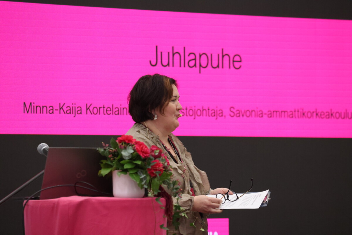 Nainen seisoo puhujakorokkeella kukkien keskellä, paperit kädessään ja puhuu. Hänen takanaan on suuri vaaleanpunainen näyttö, jossa näkyy suomenkielinen sana Juhlapuhe sekä hänen nimensä, Minna-Kaija Kortelainen, ja hänen tittelinsä sekä organisaationsa.