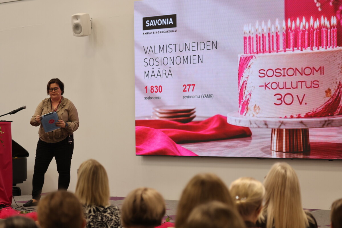 Nainen puhuu lavalla suuren näytön vieressä, jolla näkyy teksti SOSIONOMI-KOULUTUS 30 V. sekä syntymäpäiväkakku ja tilastotietoja Savonian valmistuneista. Yleisö istuu häntä vastapäätä.